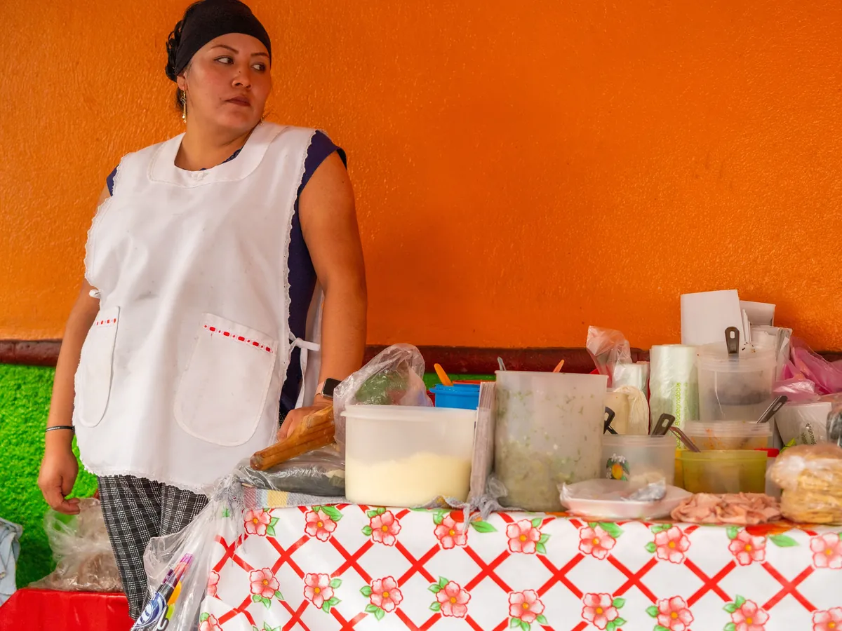 A merchant standing at her own stall, in front of an orange wall